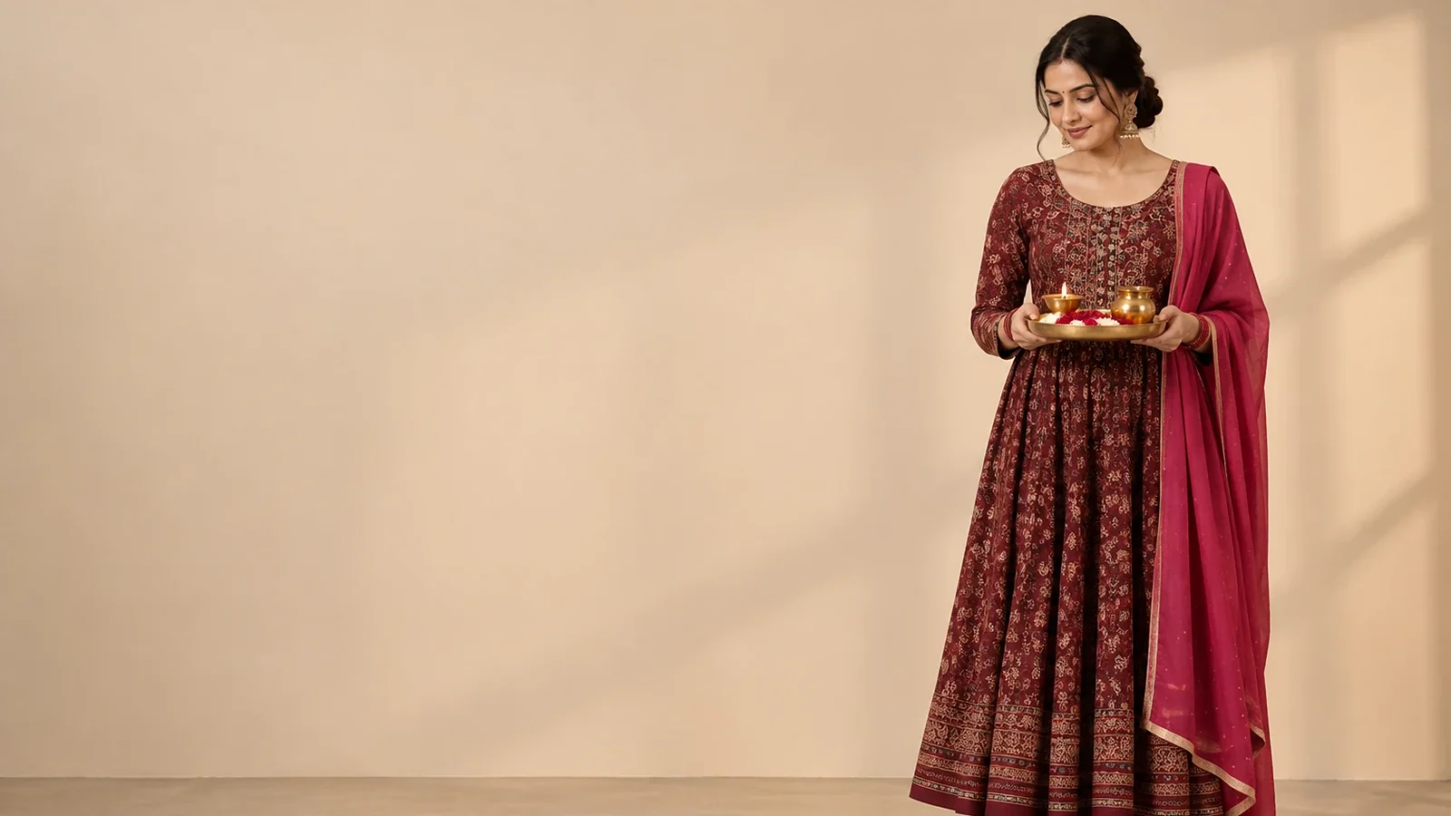 A woman in a maroon printed Anarkali kurta set with a pink dupatta, holding a brass puja thali with a lit diya