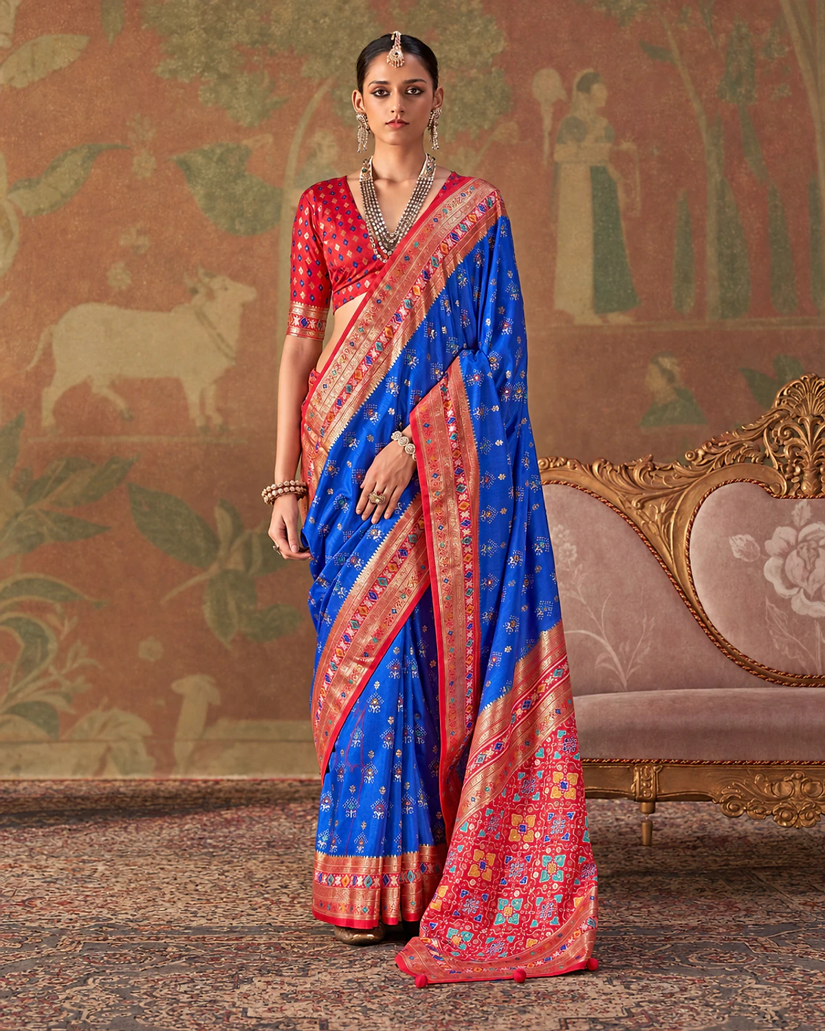 Woman wearing a royal blue Tarini Patola-style sigma silk saree with a red-pink border and patterned pallu, standing beside a carved sofa