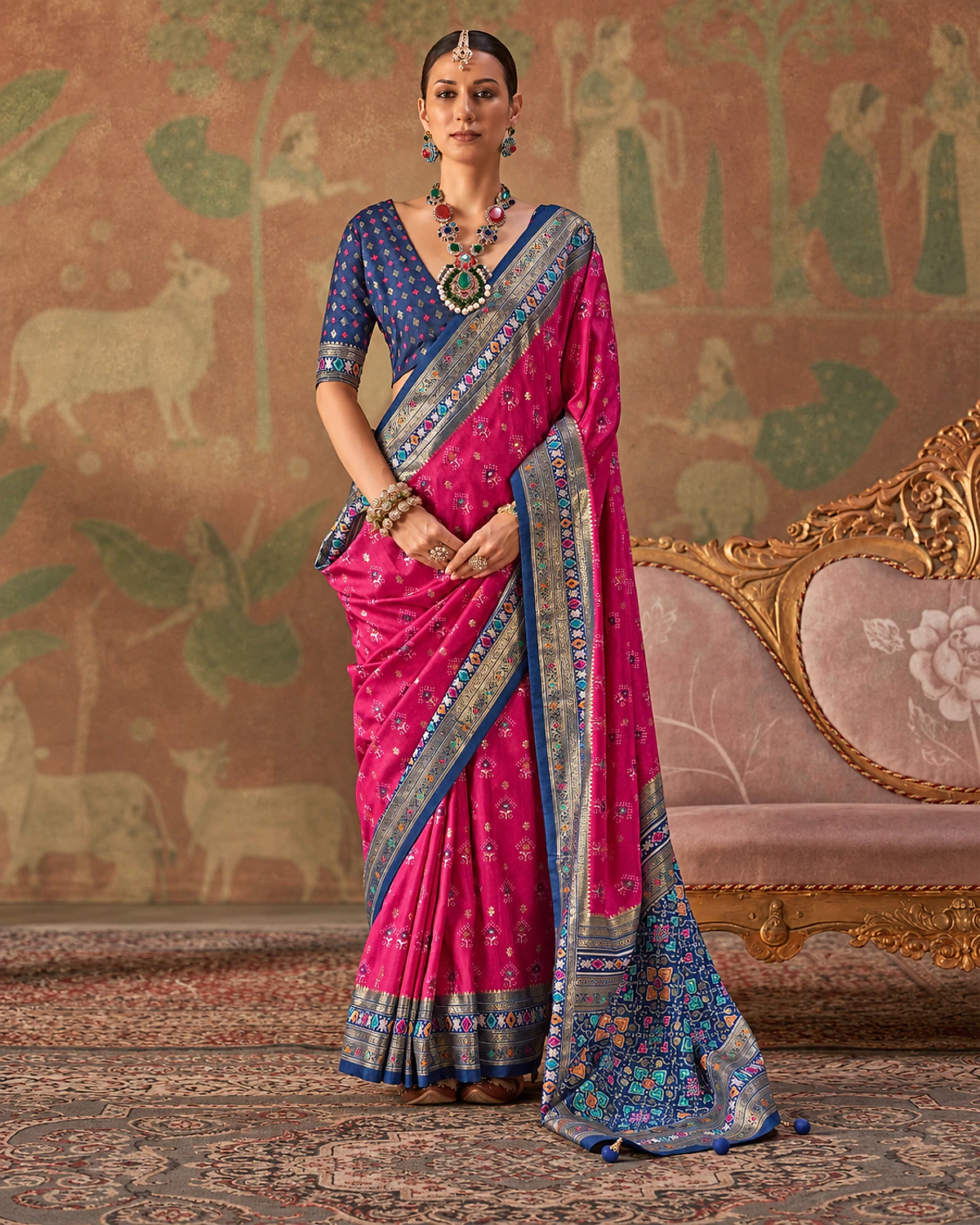 Woman wearing a magenta Tarini Patola-style sigma silk saree with a slate-blue and aqua border and patterned pallu, standing beside a carved sofa