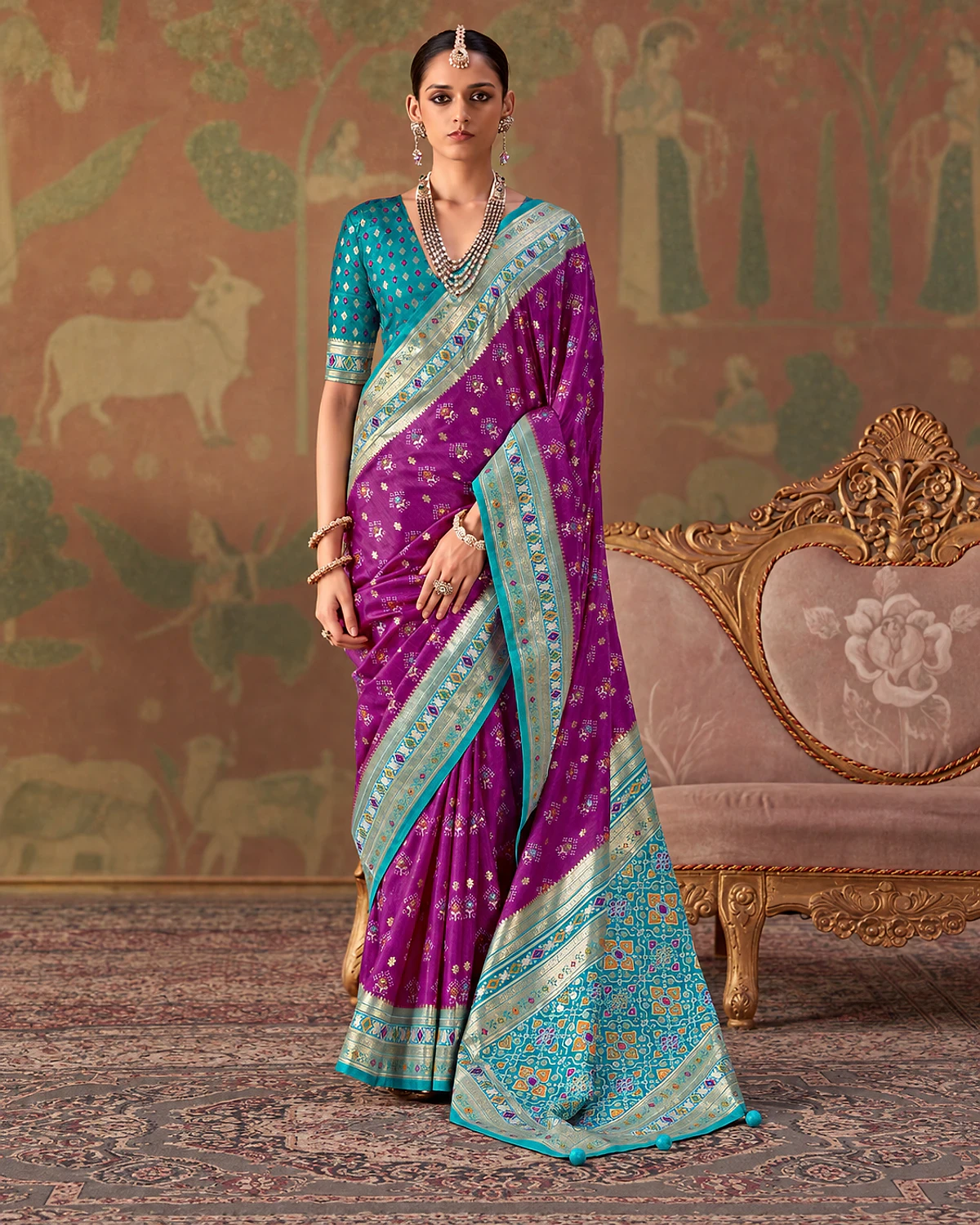 Woman wearing a purple Tarini Patola-style sigma silk saree with a turquoise and aqua border and patterned pallu, standing beside a carved sofa