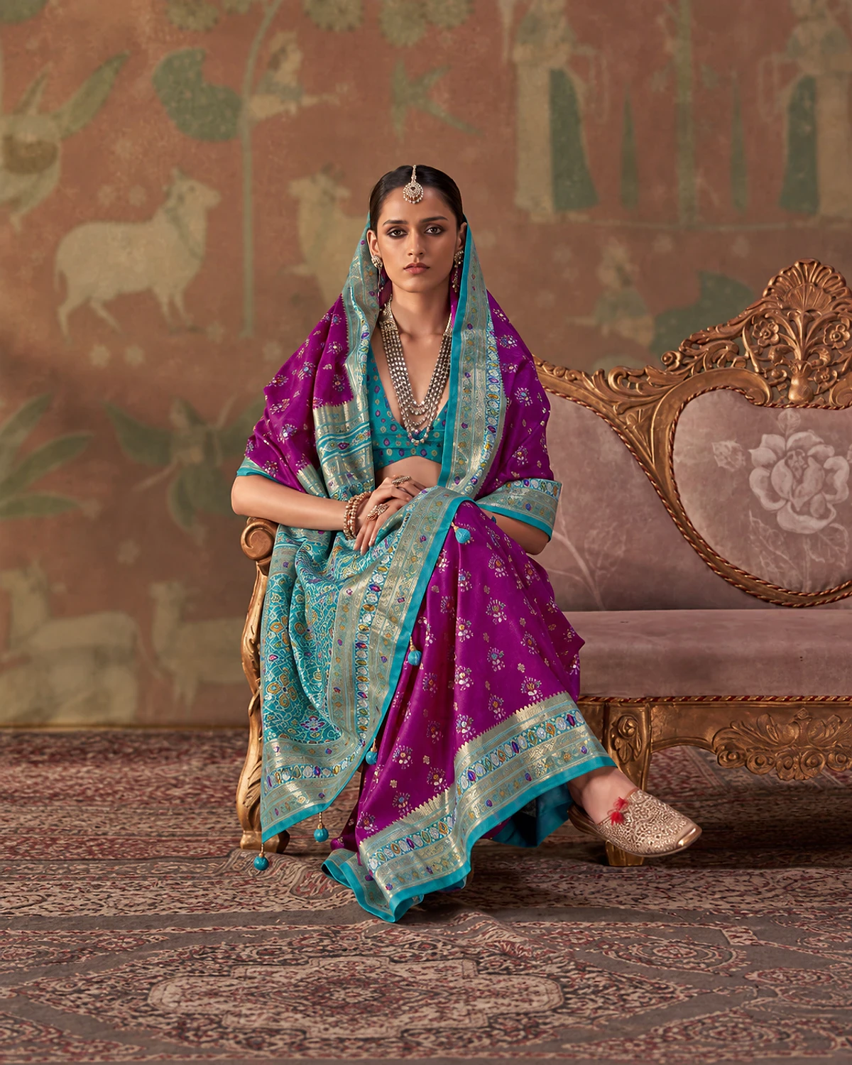 Woman seated on a carved sofa in the purple Tarini Patola-style sigma silk saree, its turquoise pallu draped over her head