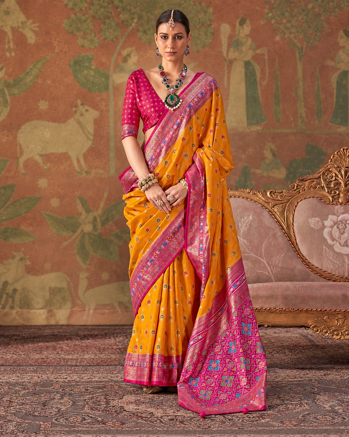 Woman wearing a mustard Tarini Patola-style sigma silk saree with a magenta-pink border and patterned pallu, standing beside a carved sofa