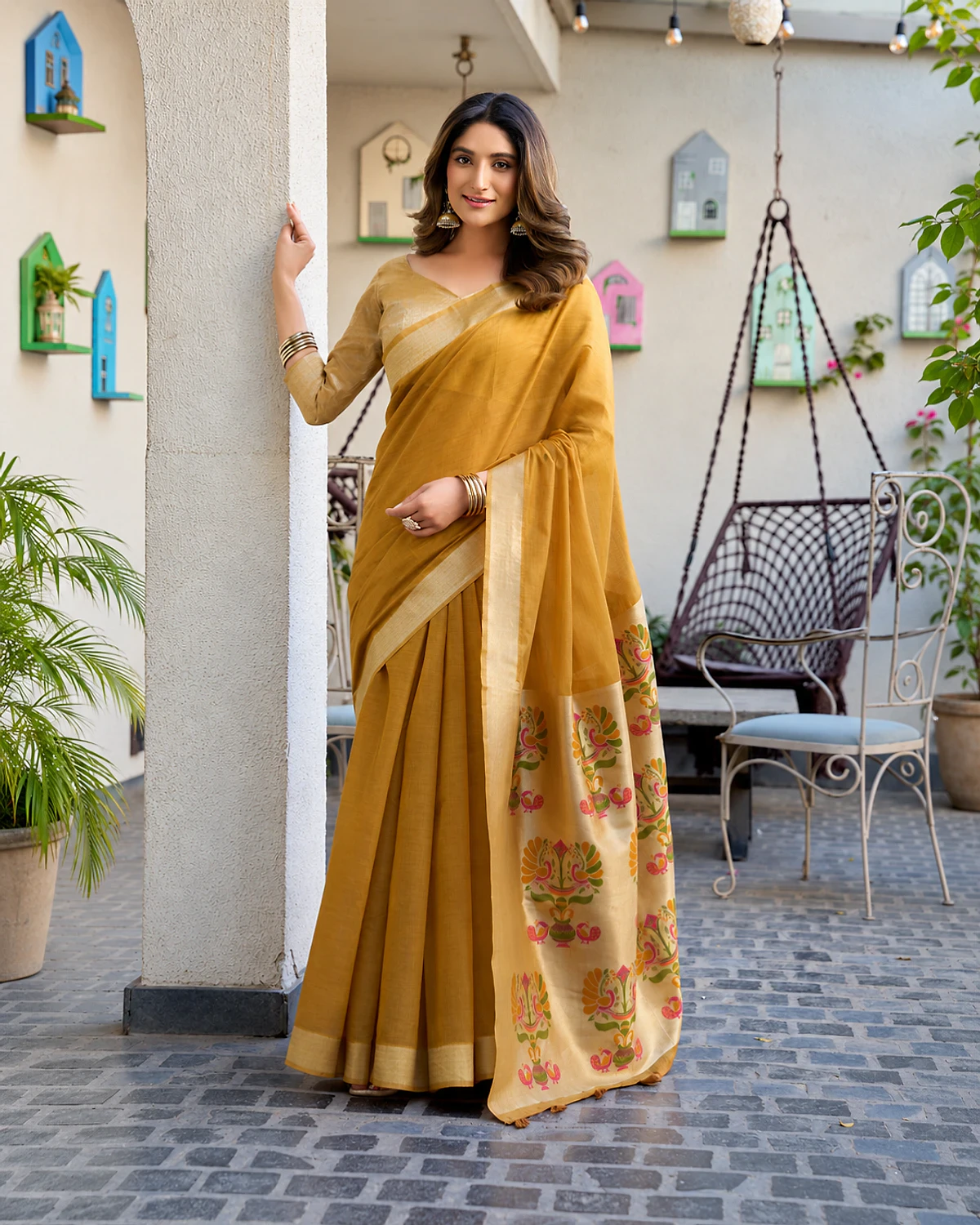 Woman wearing a mustard Surbhi cotton jacquard saree with a zari border and Paithani-style pallu, standing by a white pillar in a courtyard