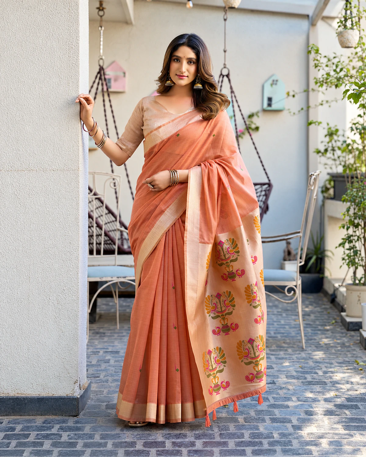 Woman wearing a peach Surbhi cotton jacquard saree with a zari border and Paithani-style pallu, standing by a white pillar in a courtyard