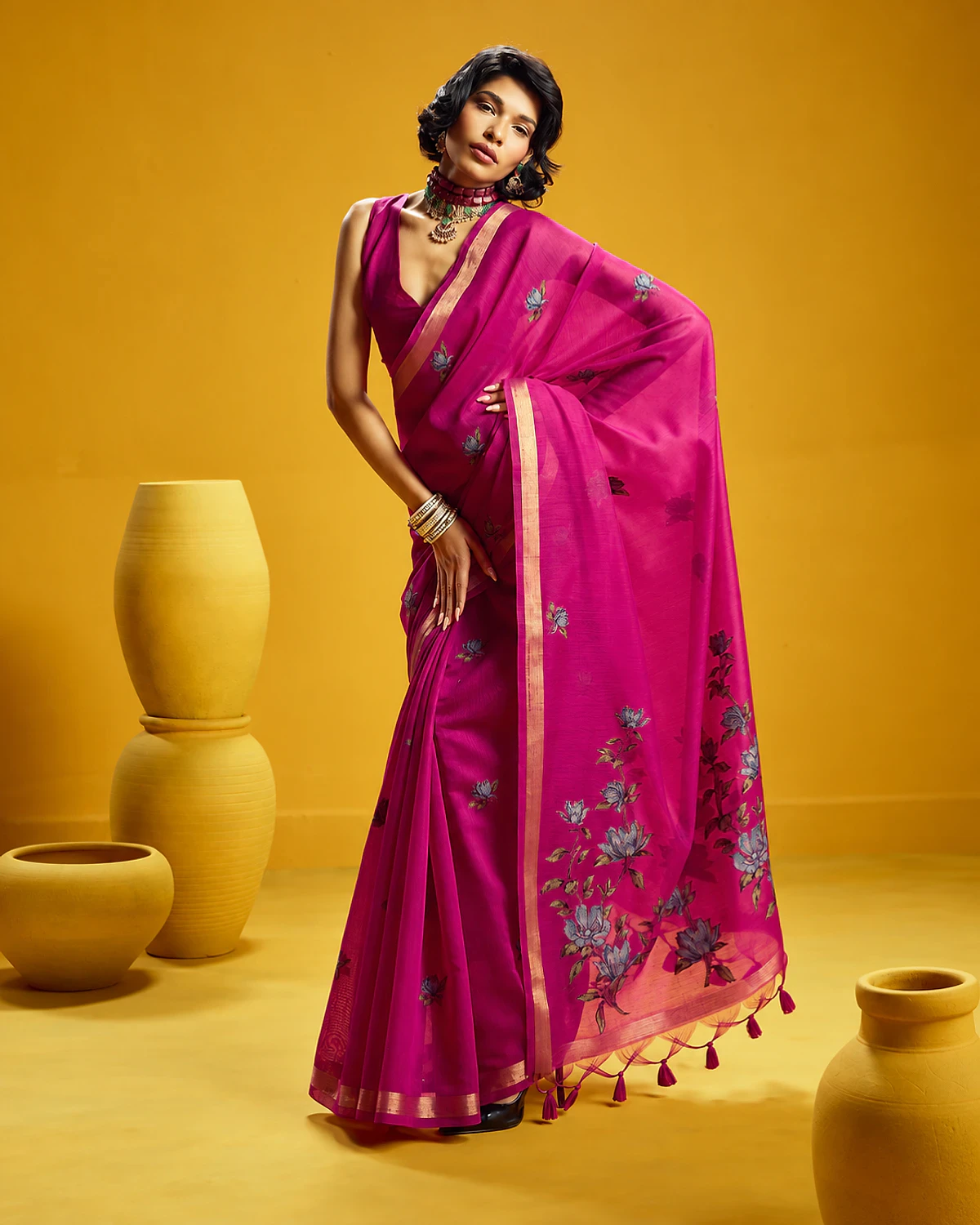 Woman wearing a magenta Noorani handloom munga cotton saree with woven flowers and a golden border, standing among mustard-yellow pots