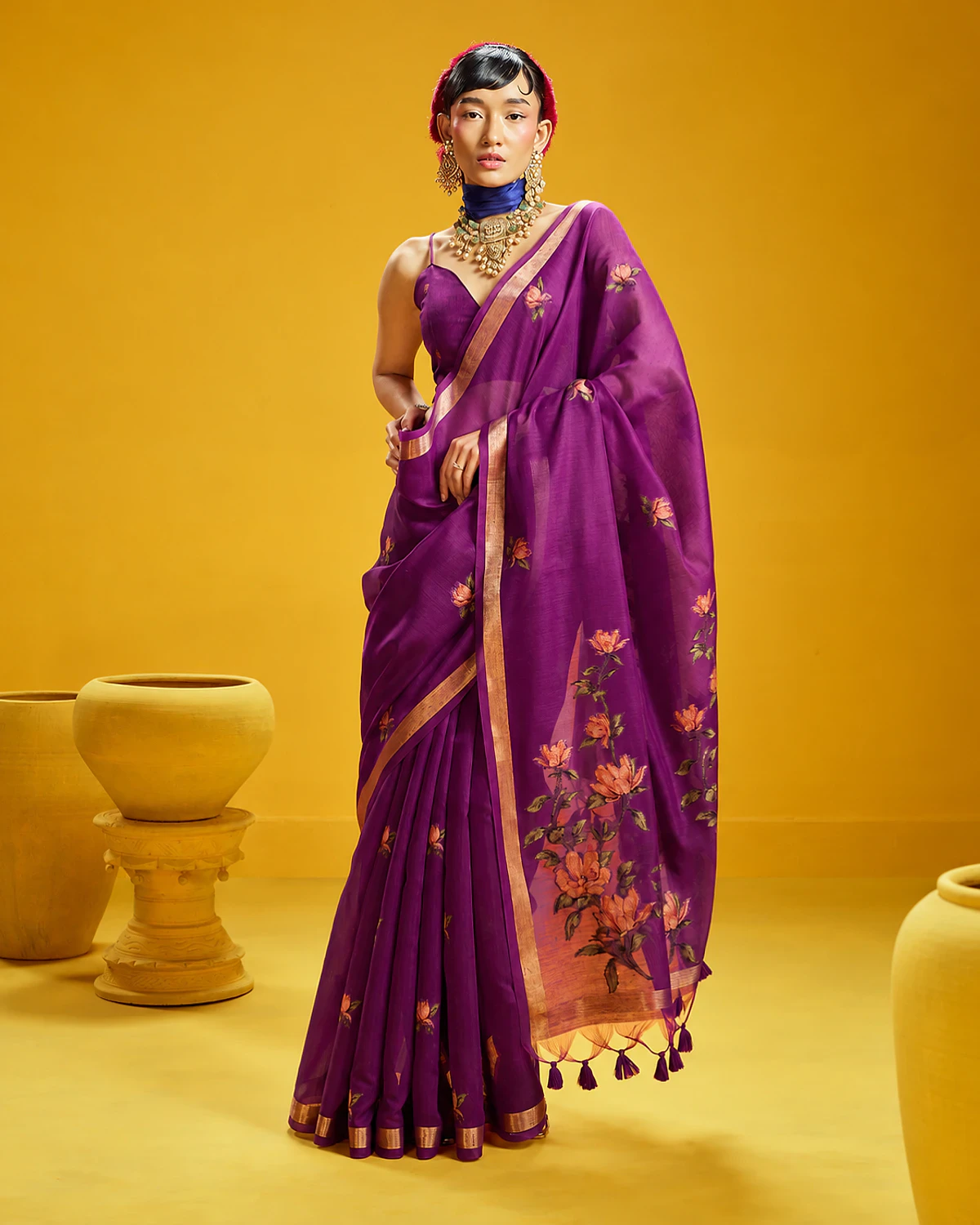 Woman wearing a purple Noorani handloom munga cotton saree with woven flowers and a golden border, standing among mustard-yellow pots