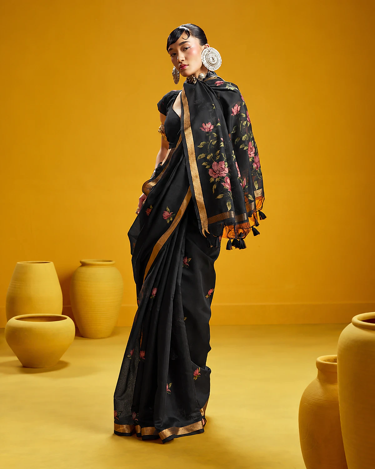 Woman wearing a black Noorani handloom munga cotton saree with woven flowers and a golden border, standing among mustard-yellow pots
