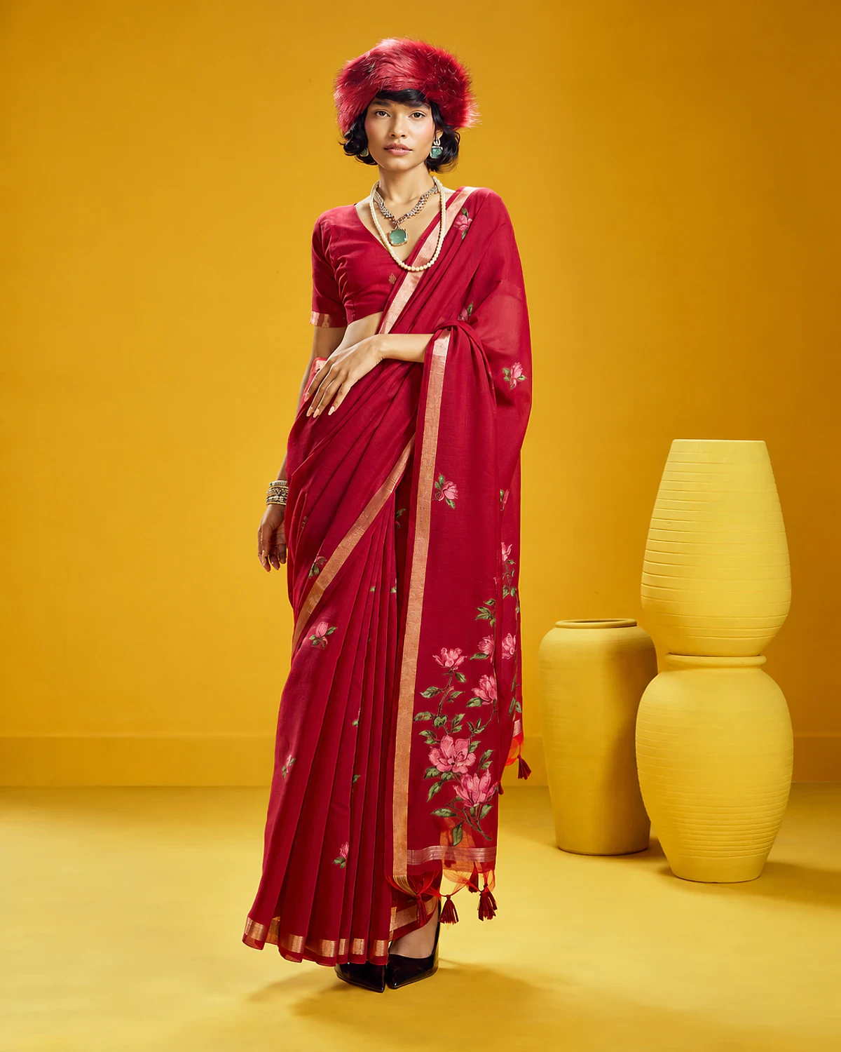 Woman wearing a crimson Noorani handloom munga cotton saree with woven flowers and a golden border, standing among mustard-yellow pots