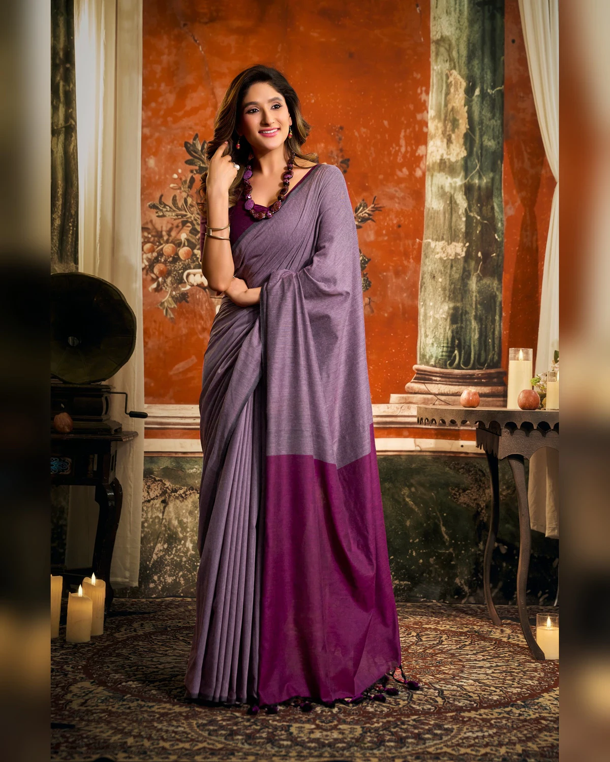 Woman wearing a lilac grey mal cotton saree with a wide purple pallu, standing in a room with a painted wall and candles