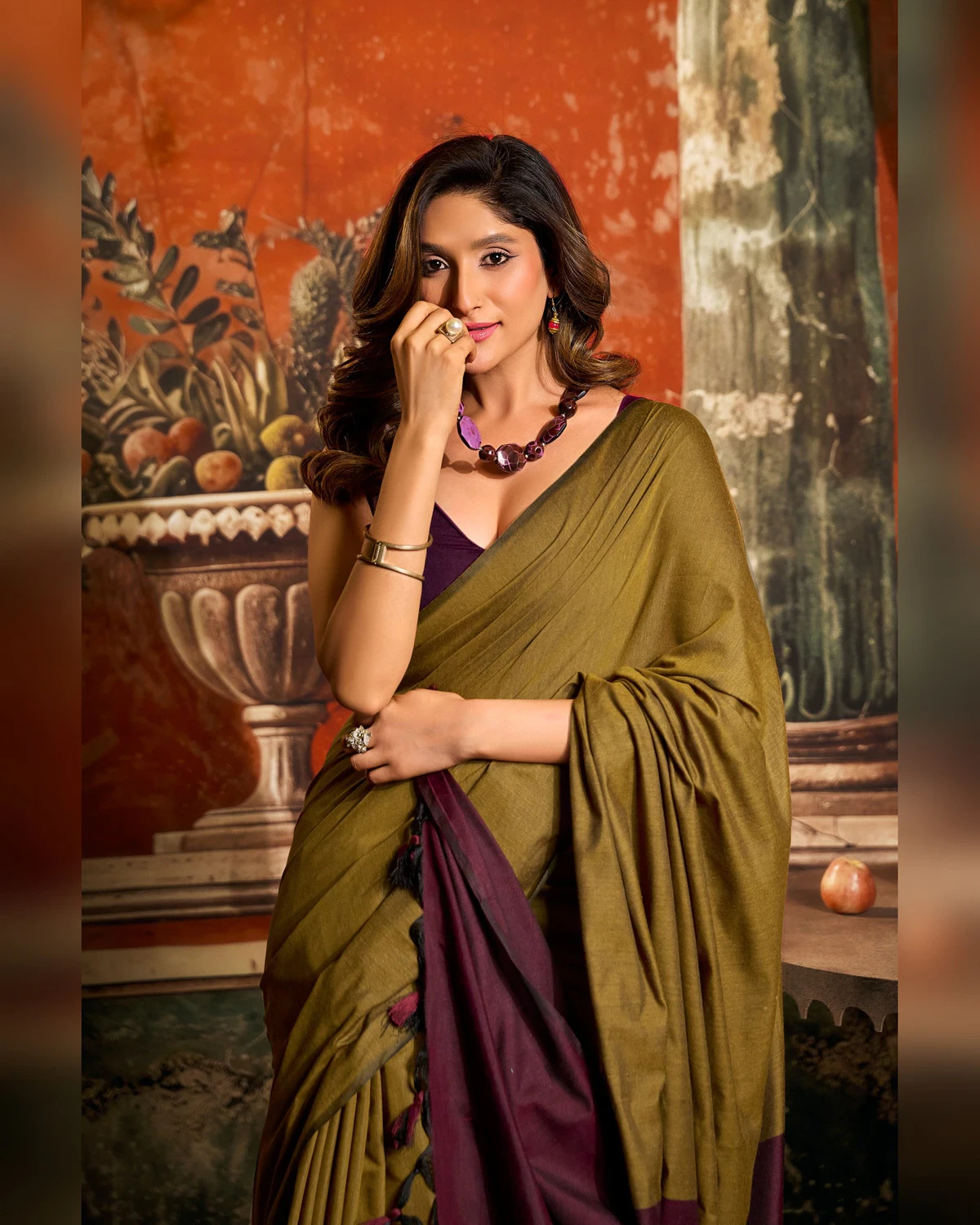 Woman wearing an olive green mal cotton saree with a maroon pallu, seen from the waist up in a room with a painted wall