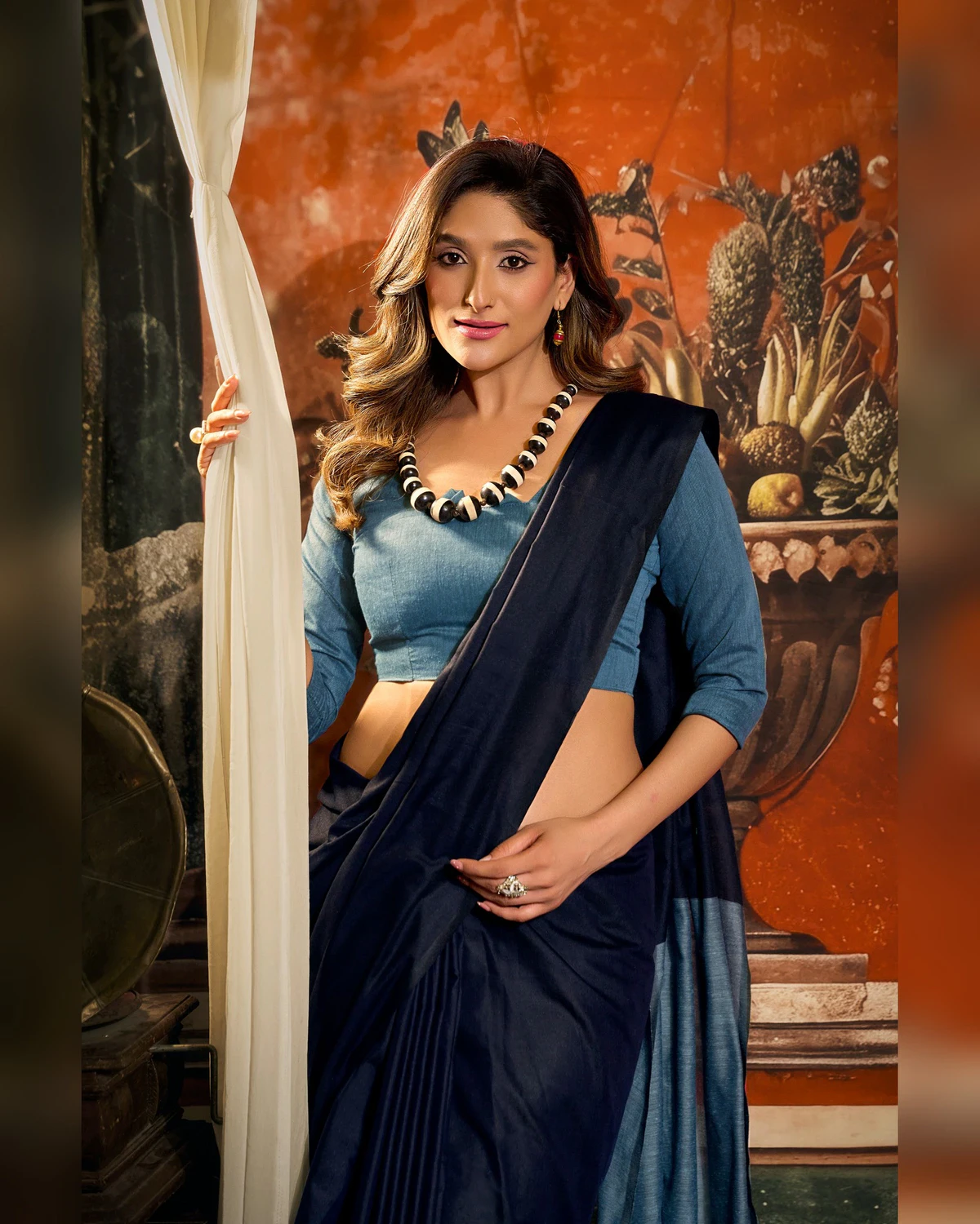Woman in the navy blue mal cotton saree and a powder blue blouse, seen from the waist up beside a curtain