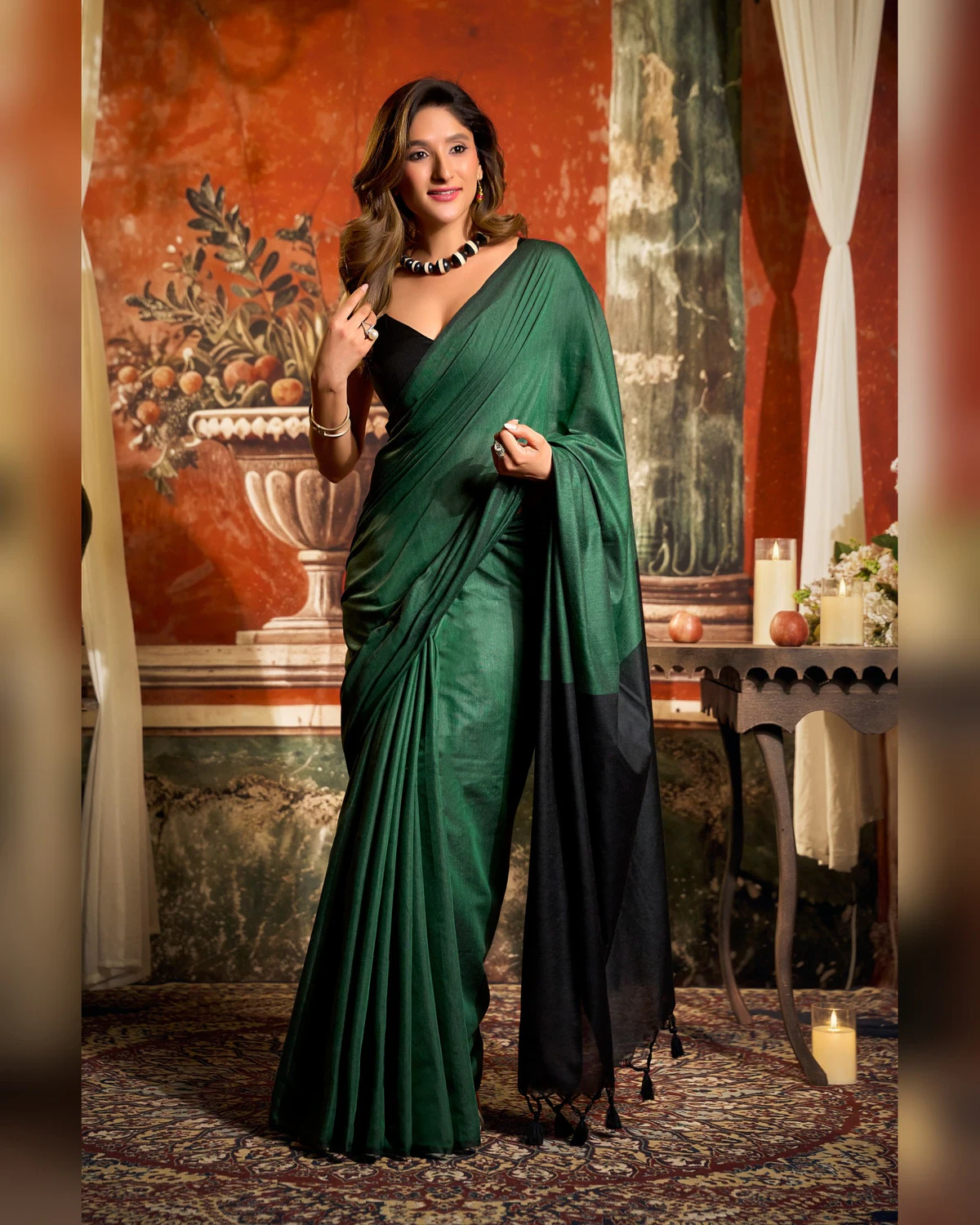 Woman wearing a bottle green mal cotton saree with a black pallu and black blouse, standing in a room with a painted wall