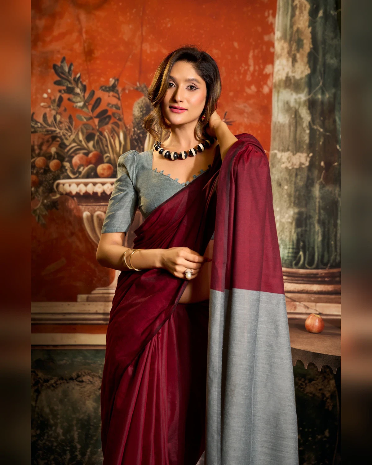 Woman in the maroon mal cotton saree holding its grey pallu, seen from the waist up