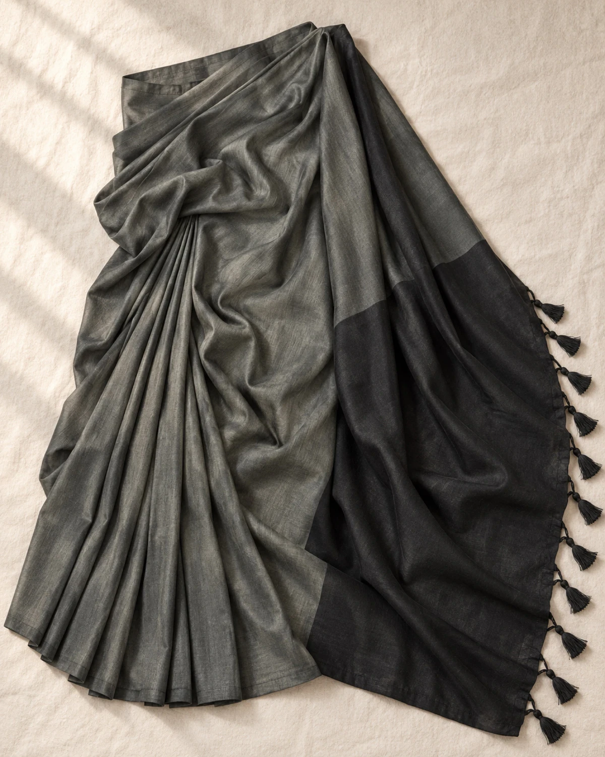 The charcoal grey mal cotton saree laid flat, showing its wide black pallu and tassels