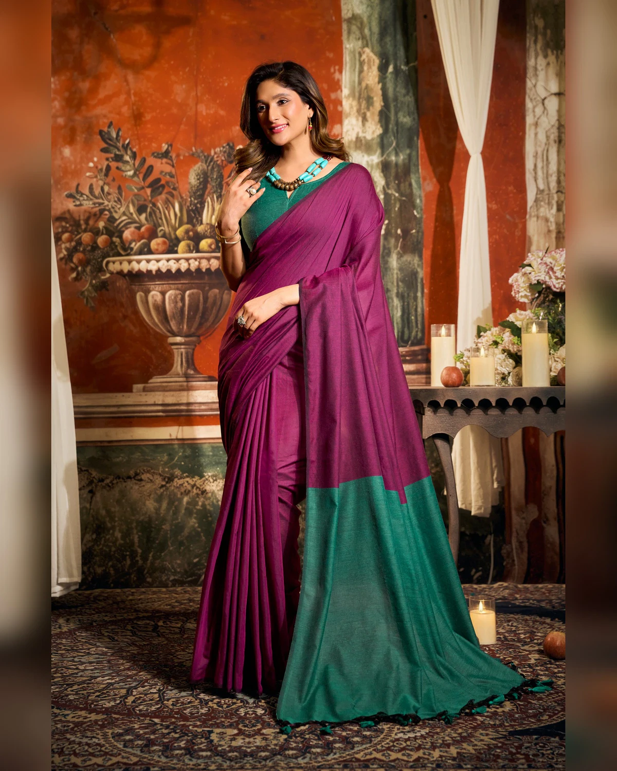 Woman wearing a magenta mal cotton saree with a teal-green pallu and blouse, standing in a room with a painted wall