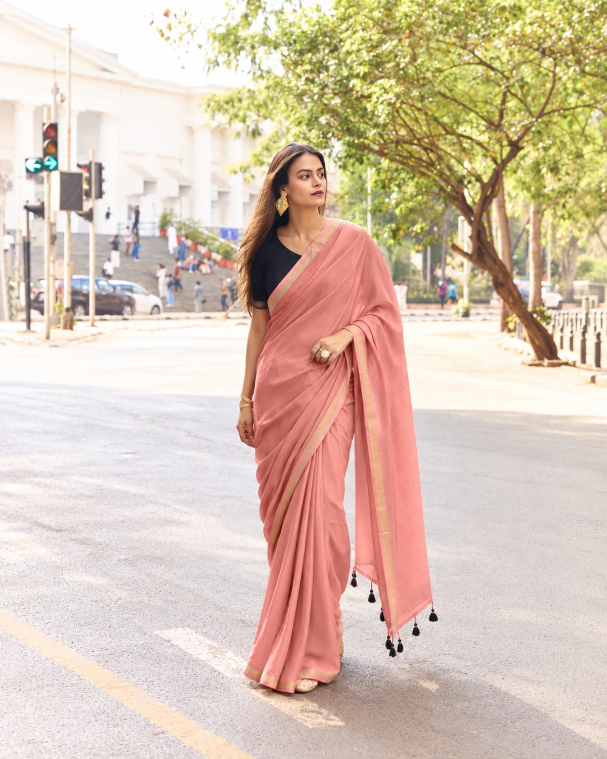 Woman standing on a tree-lined street in the dusty pink mul mul cotton saree, its tasselled pallu over her arm