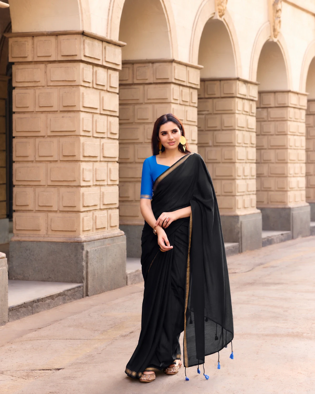 Woman wearing a black mul mul cotton saree with a golden border and a royal blue blouse, standing under stone arches