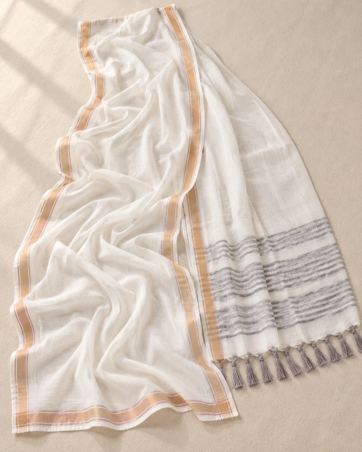 The white mul mul cotton saree laid flat, showing its golden border and grey-striped pallu with tassels