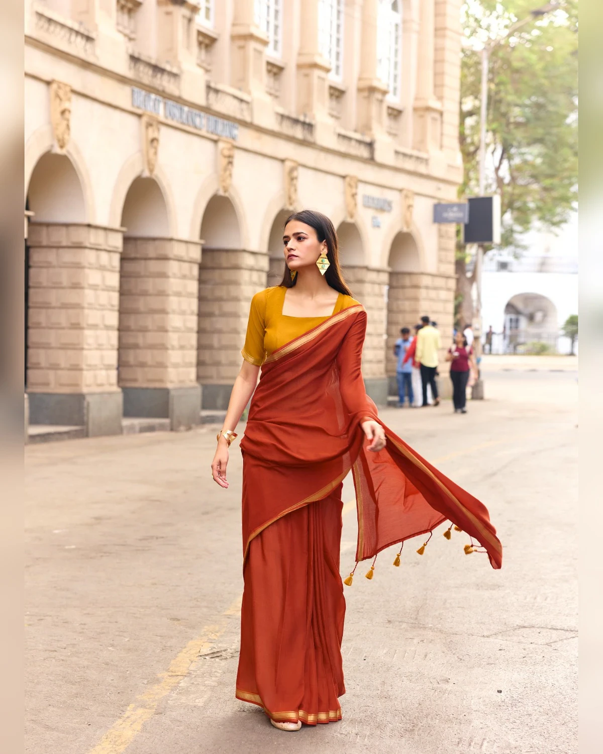 Woman walking down a street in a rust mul mul cotton saree with a mustard blouse, its pallu lifting in the breeze