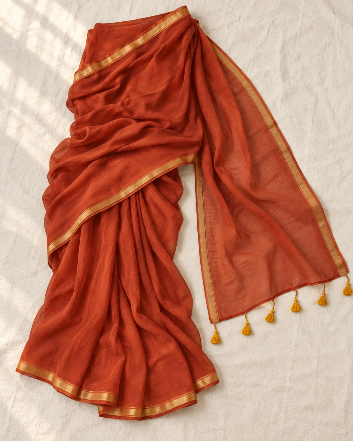 The rust mul mul cotton saree laid flat, showing its golden border and mustard tassels