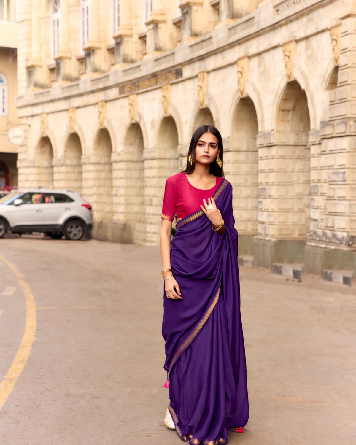 Woman wearing a purple mul mul cotton saree with a golden border and a pink blouse, standing on a street with arches behind