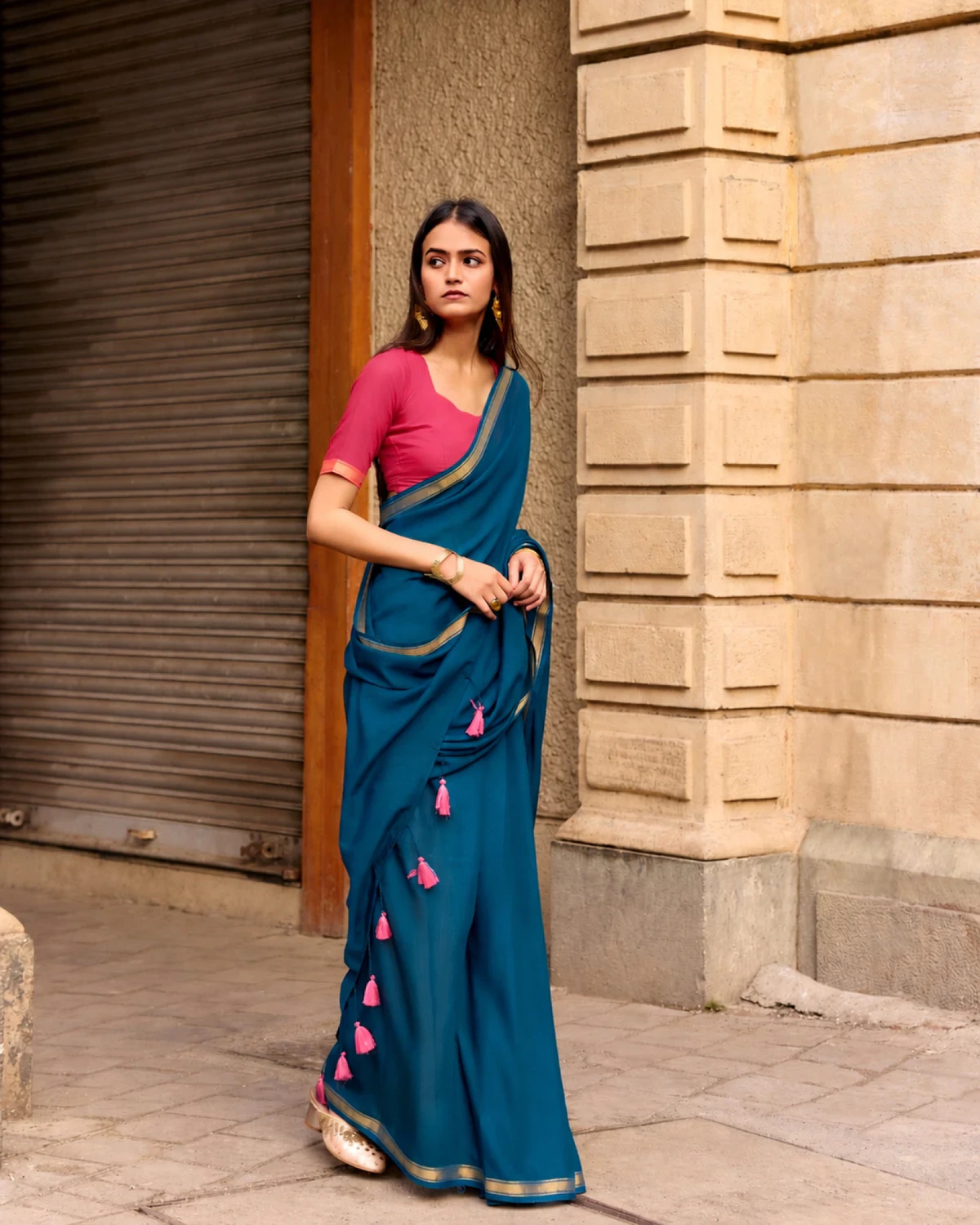 Woman wearing a teal blue mul mul cotton saree with a golden border and a pink blouse, standing by a shop shutter