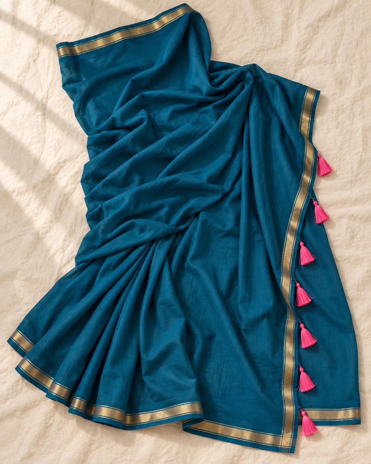 The teal blue mul mul cotton saree laid flat, showing its golden border and pink tassels