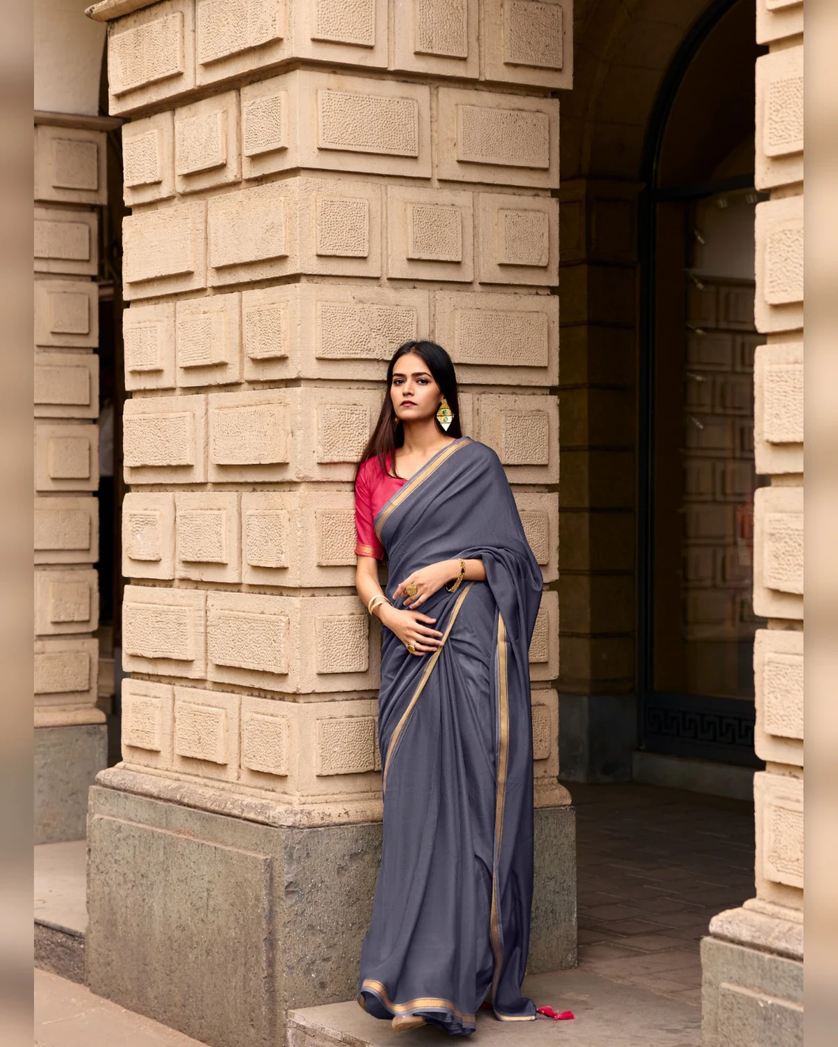 Woman in a grey mul mul cotton saree with a golden border and a pink blouse, leaning against a stone pillar