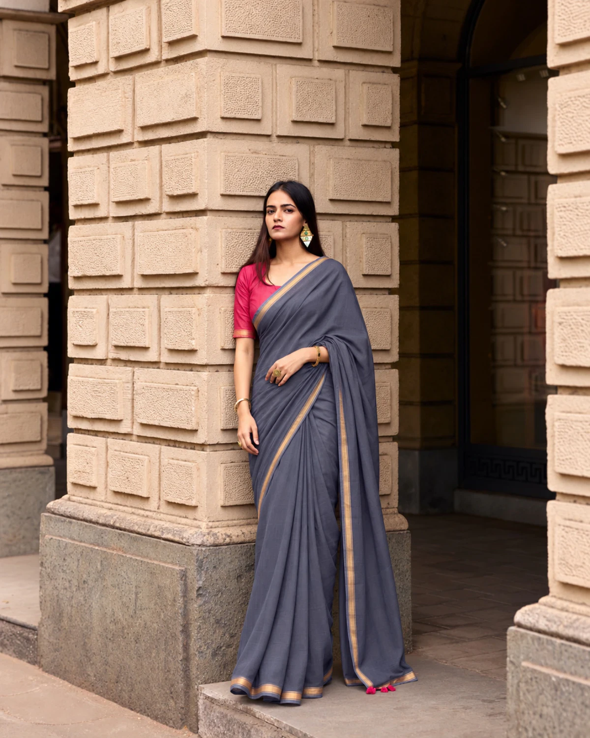 Woman standing in the grey mul mul cotton saree by a stone pillar, its pallu falling over her arm