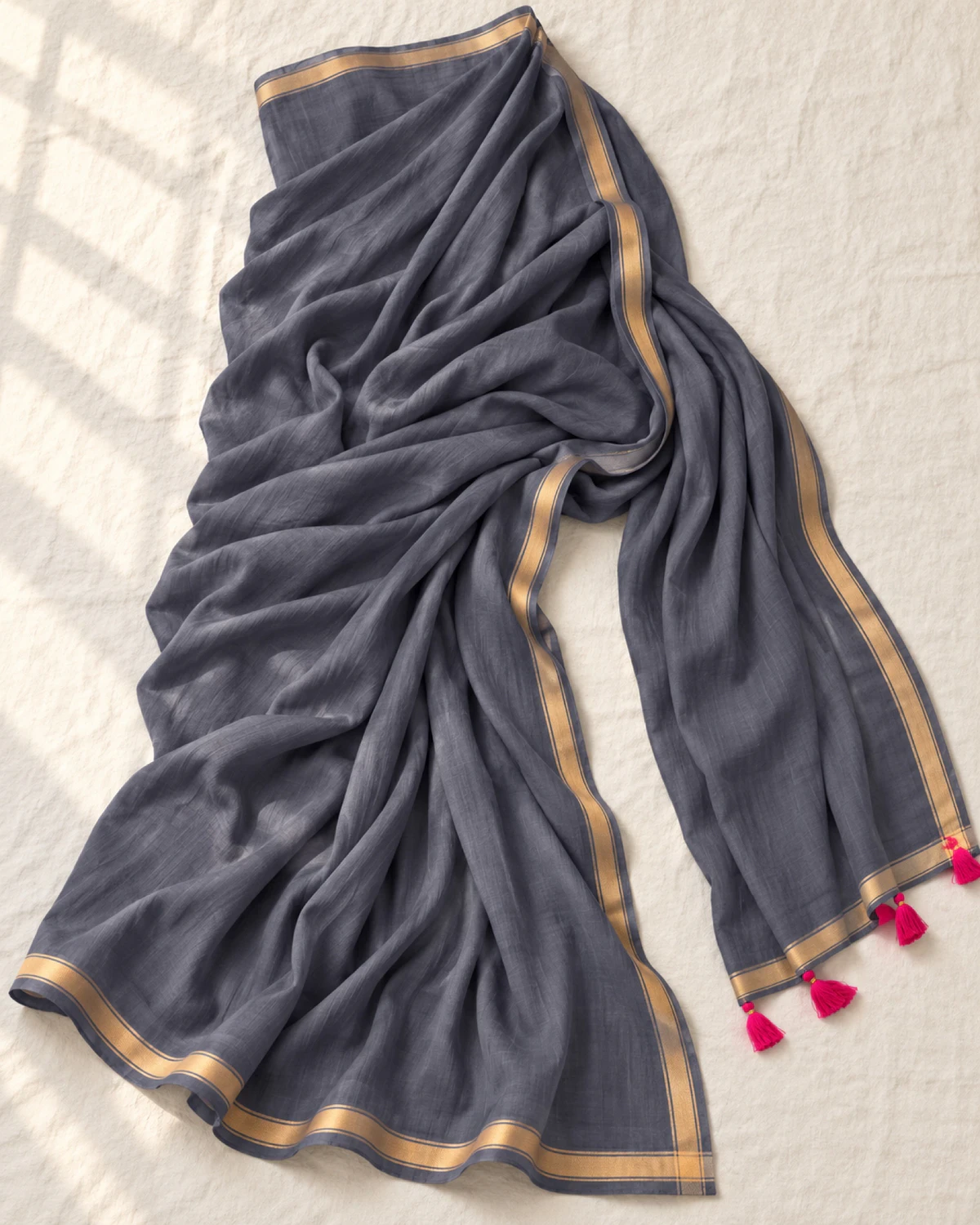 The grey mul mul cotton saree laid flat, showing its golden border and pink tassels