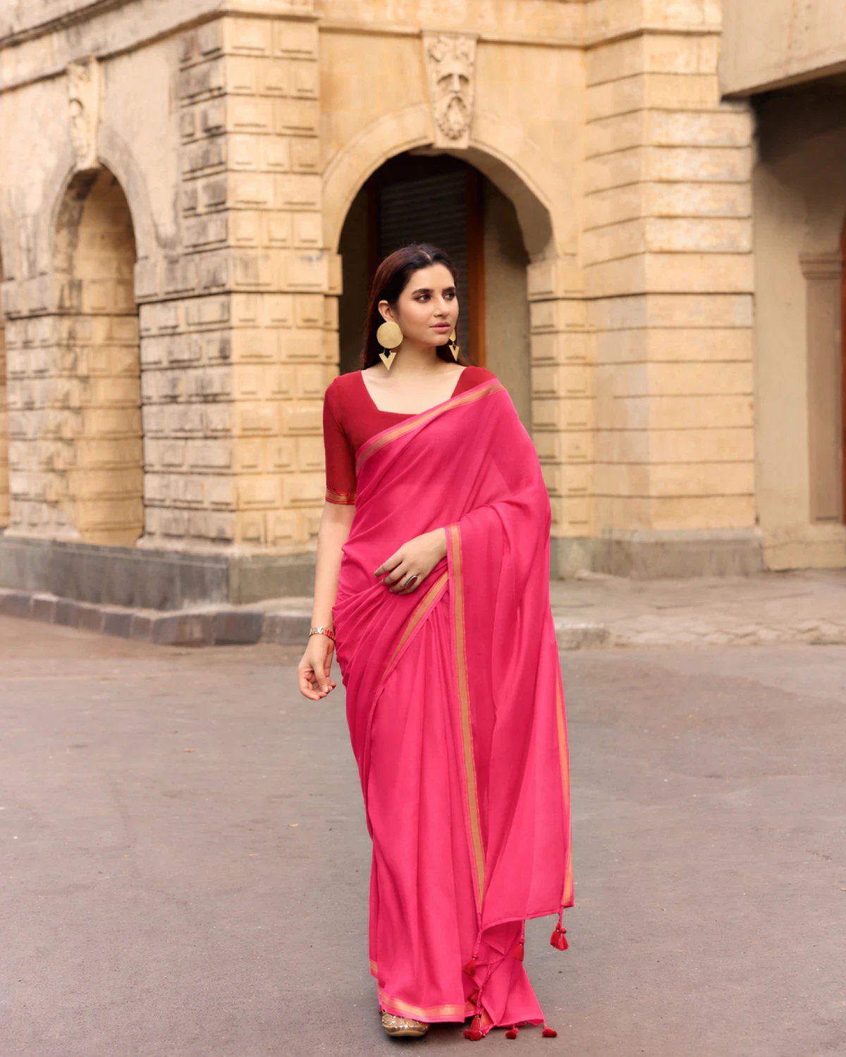 Woman wearing a rani pink mul mul cotton saree with a golden border and a deep red blouse, standing on a street with arches behind
