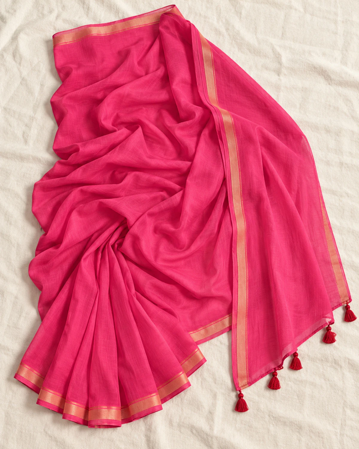 The rani pink mul mul cotton saree laid flat, showing its golden border and red tassels