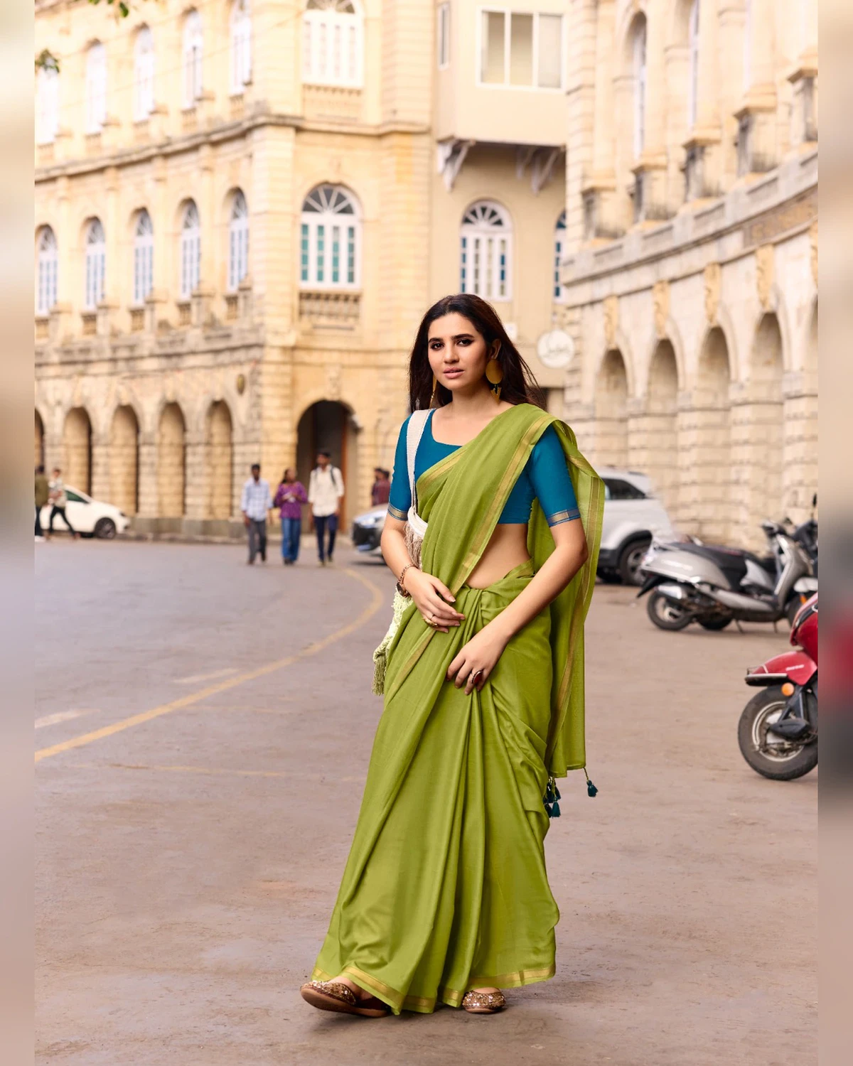 Woman walking down a city street in an olive green mul mul cotton saree with a golden border and a teal blue blouse