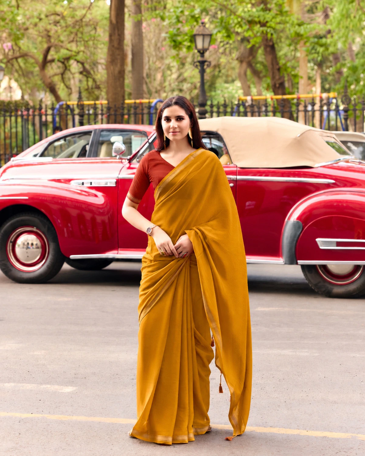 Woman wearing a mustard yellow mul mul cotton saree with a golden border and a rust blouse, standing by a red vintage car