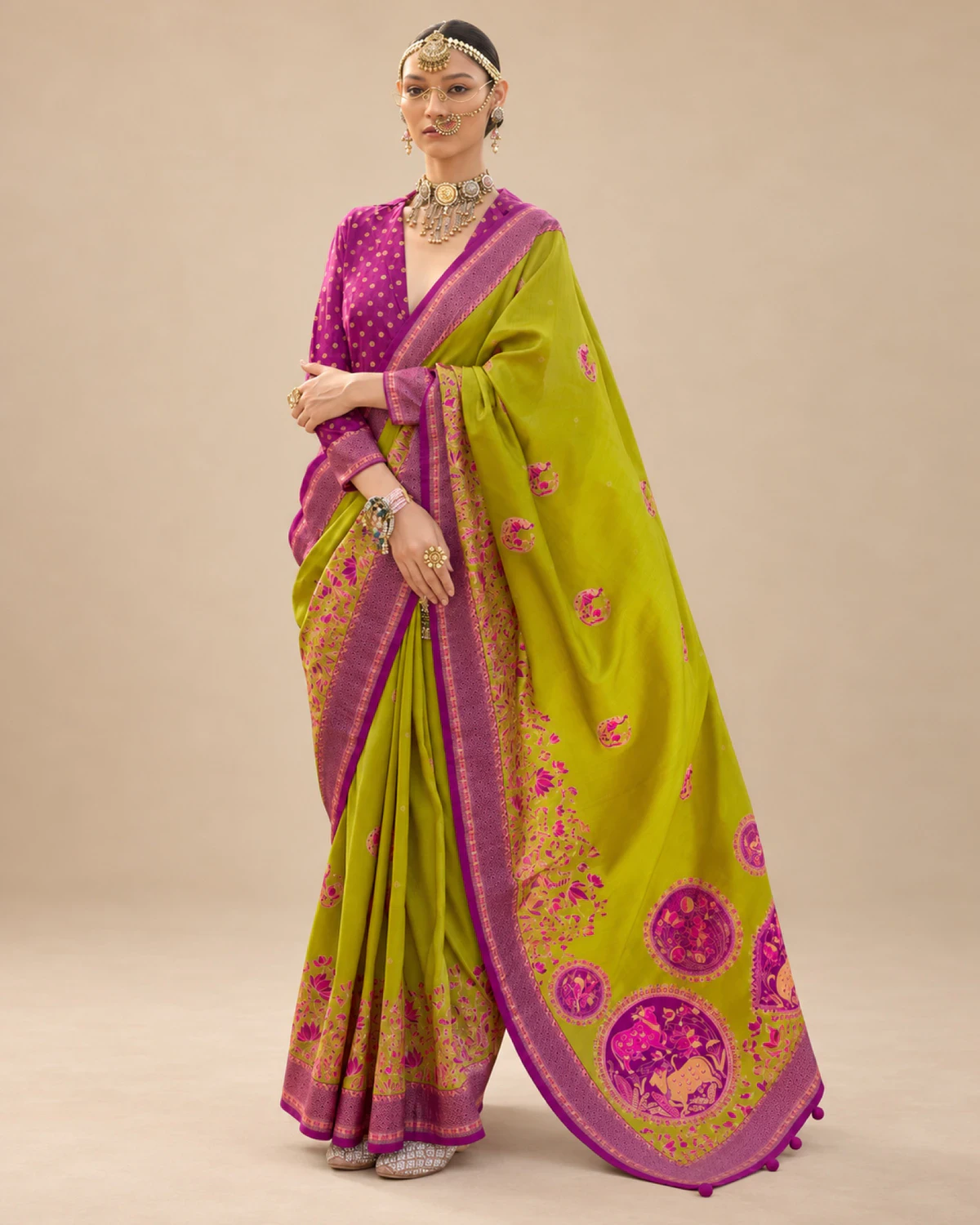 Woman wearing a lime green printed sigma silk saree with a purple border and medallion pallu, standing against a beige wall