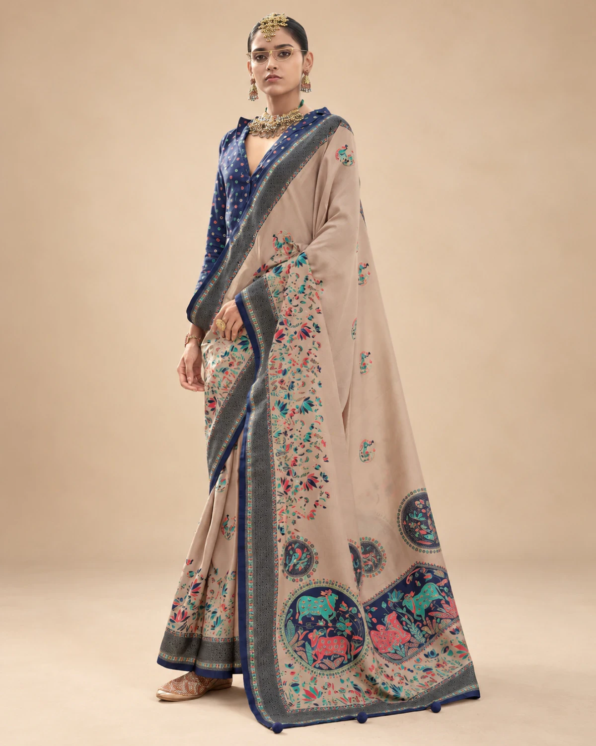 Woman wearing a beige printed sigma silk saree with a navy blue border and medallion pallu, standing against a beige wall