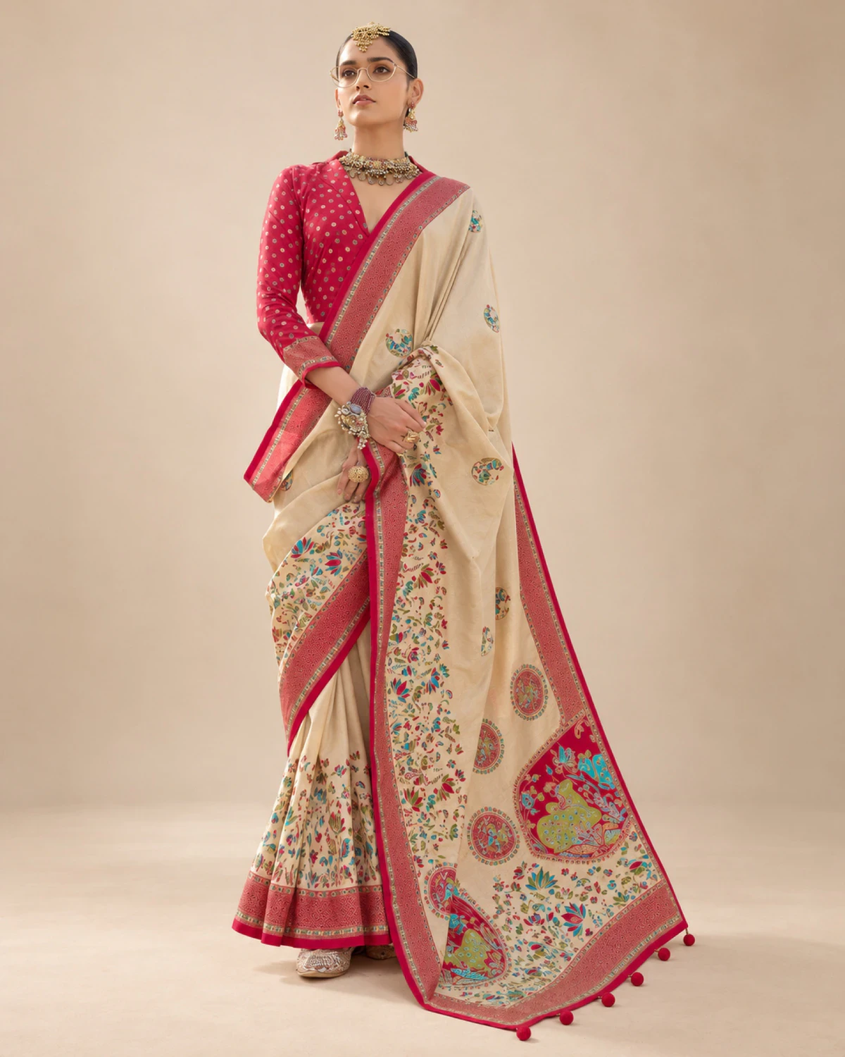 Woman wearing a cream printed sigma silk saree with a red border and medallion pallu, standing against a beige wall