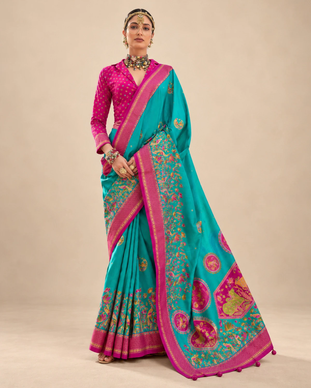 Woman wearing a teal printed sigma silk saree with a magenta pink border and medallion pallu, standing against a beige wall