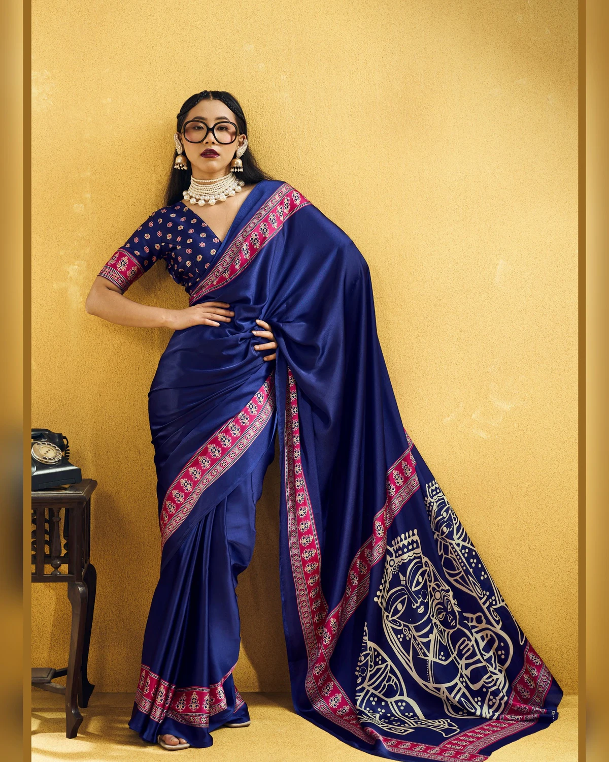 Woman wearing a navy blue modal satin saree with a rani pink Ajrakh border and line-art pallu, against a mustard wall