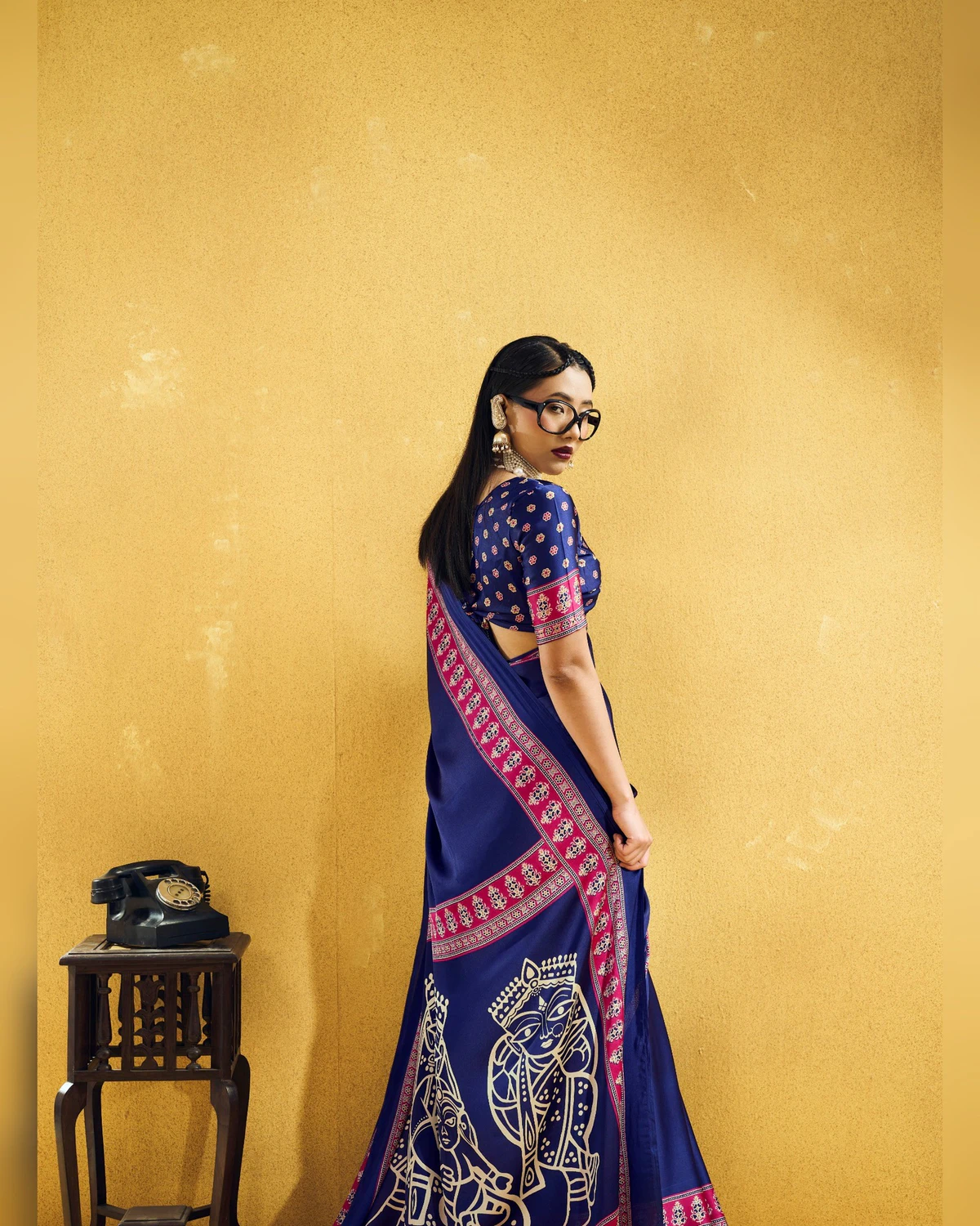 Woman in the navy blue modal satin saree seen from the back, its pallu of cream line-drawn figures falling behind her