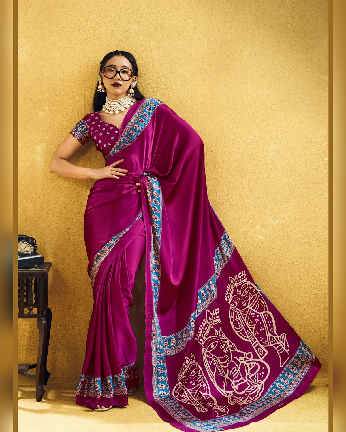 Woman wearing a rani pink modal satin saree with a blue Ajrakh border and line-art pallu, against a mustard wall