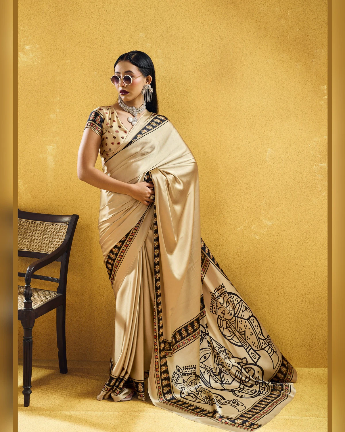 Woman wearing a beige modal satin saree with a black Ajrakh border and line-art pallu, against a mustard wall