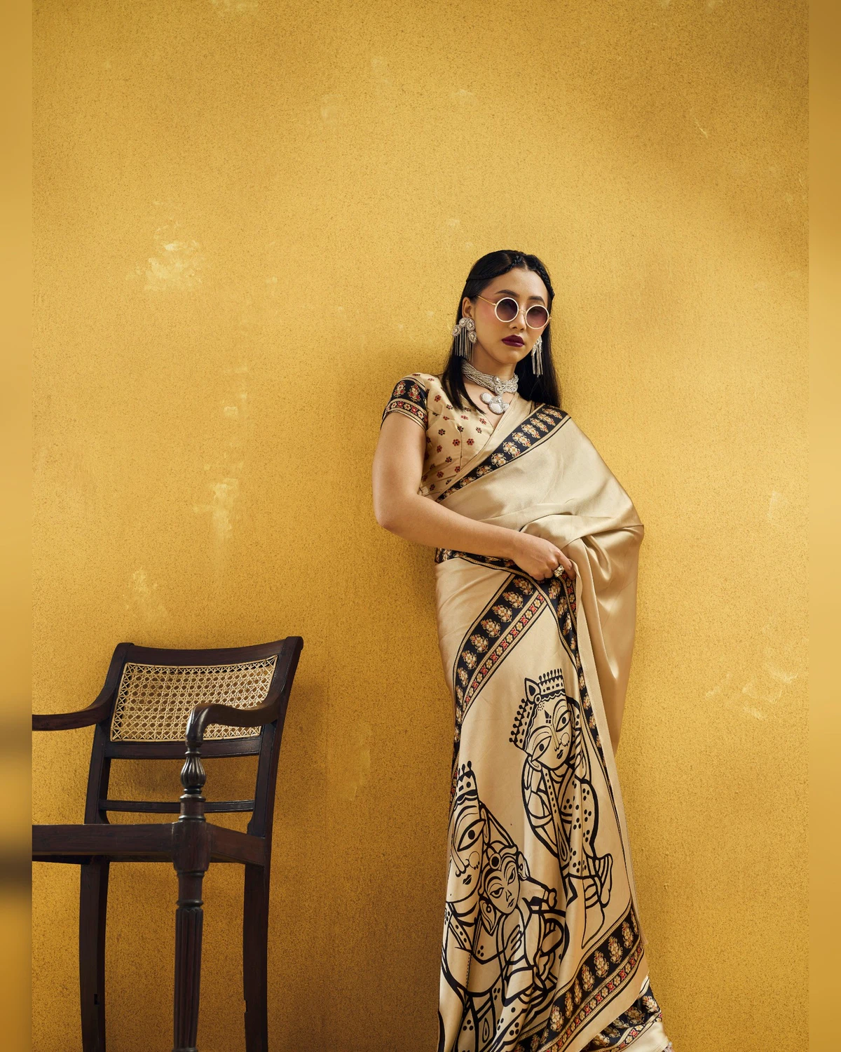 Woman in the beige modal satin saree leaning on a mustard wall, its pallu of black figures in front