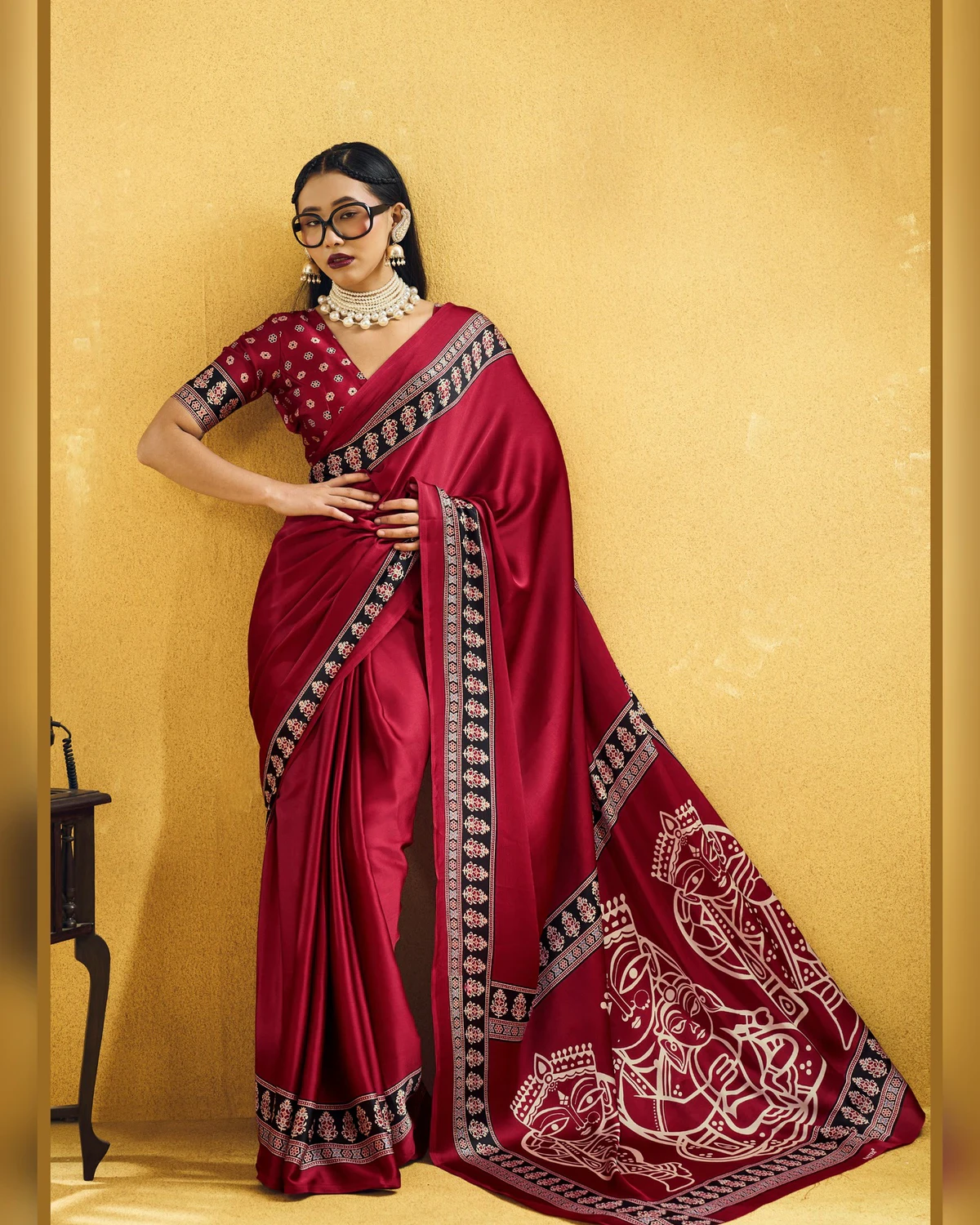 Woman wearing a red modal satin saree with a black Ajrakh border and line-art pallu, against a mustard wall