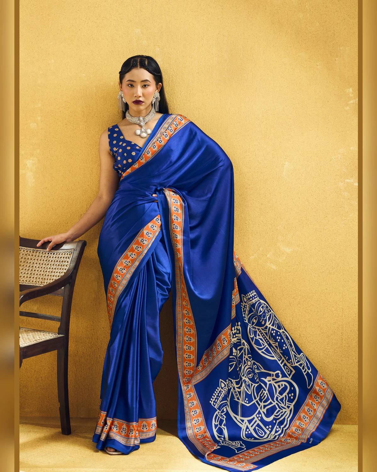 Woman wearing a royal blue modal satin saree with a orange Ajrakh border and line-art pallu, against a mustard wall