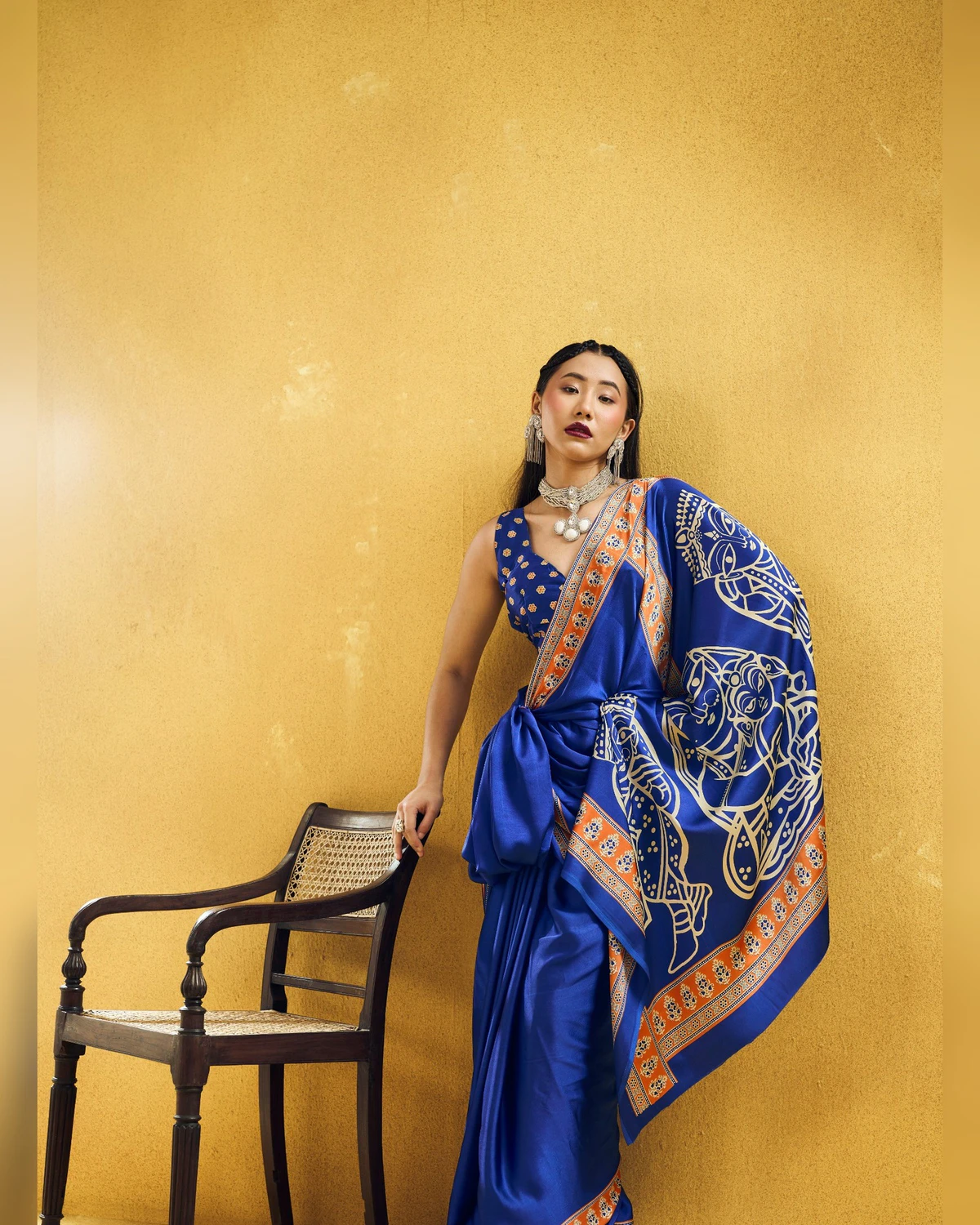Woman in the royal blue saree with its pallu draped over one arm, beside a cane chair
