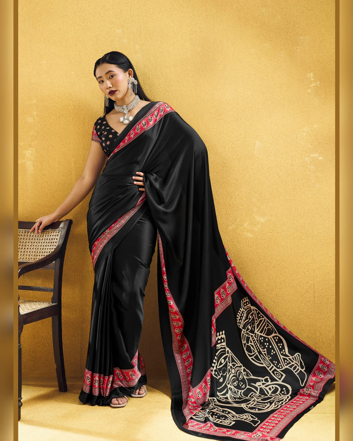 Woman wearing a black modal satin saree with a red Ajrakh border and line-art pallu, against a mustard wall