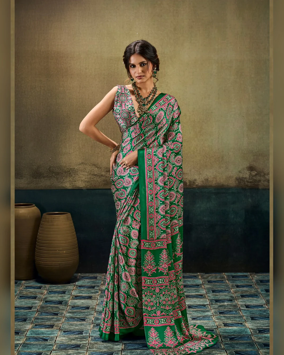 Woman wearing a emerald green all-over Ajrakh print modal satin saree, standing by clay pots on a blue tiled floor