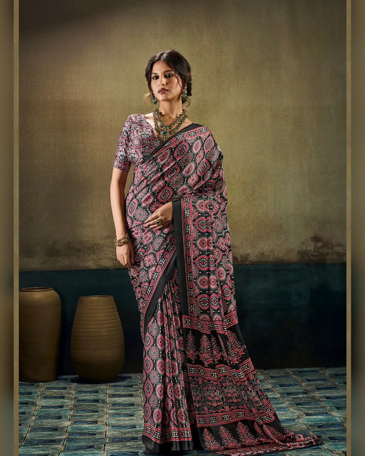 Woman wearing a black all-over Ajrakh print modal satin saree, standing by clay pots on a blue tiled floor