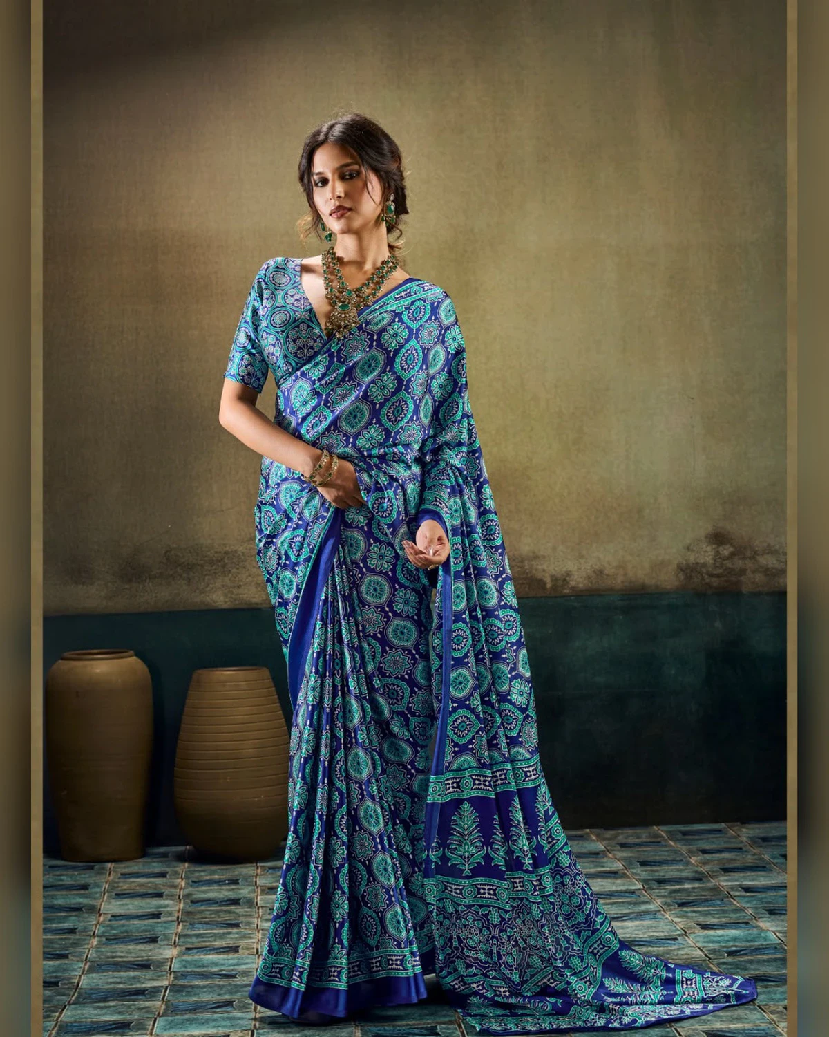 Woman wearing a blue all-over Ajrakh print modal satin saree, standing by clay pots on a blue tiled floor